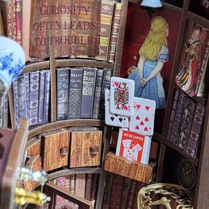 Puede incluir: Diorama de estanter&iacute;a en miniatura con tem&aacute;tica literaria. Presenta libros diminutos, naipes y una imagen enmarcada de Alicia en el Pa&iacute;s de las Maravillas. El texto "CURIOSITY OFTEN LEADS TO TROUBLE" es visible en un libro. El diorama es de madera.