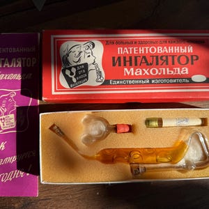 Puede incluir: Un inhalador de vidrio vintage en una caja rectangular con tapa roja y texto en ruso. El conjunto incluye una boquilla de vidrio transparente, una pequeña pipa de vidrio ámbar con hendiduras circulares y un frasco pequeño con tapón.
