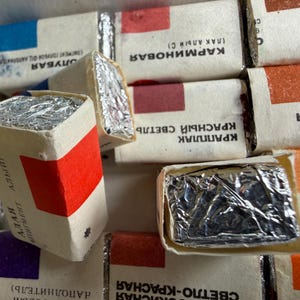 May include: A collection of rectangular watercolor paint pans wrapped in white paper with silver foil ends. Each pan features a colored square indicating the pigment shade and Cyrillic text labels. The arrangement shows multiple individual paint blocks.