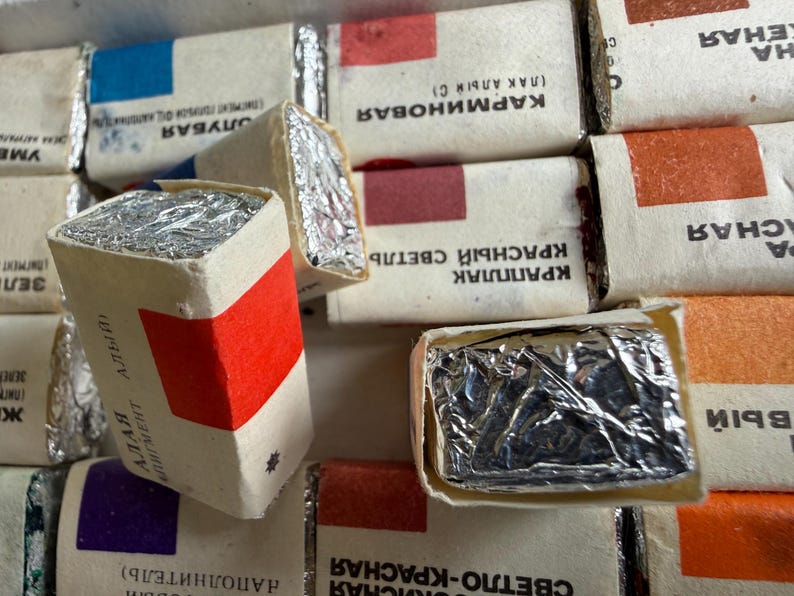 May include: A collection of rectangular watercolor paint pans wrapped in white paper with silver foil ends. Each pan features a colored square indicating the pigment shade and Cyrillic text labels. The arrangement shows multiple individual paint blocks.