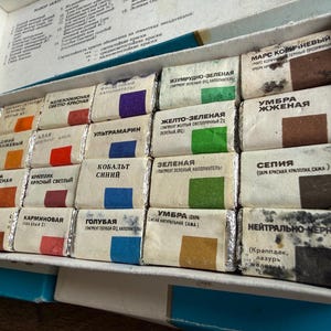 May include: A rectangular white box containing twenty-four individual watercolor paint pans wrapped in paper. Each pan features a color swatch and Russian text labels. The set includes various shades of yellow, orange, red, blue, green, brown, and black.