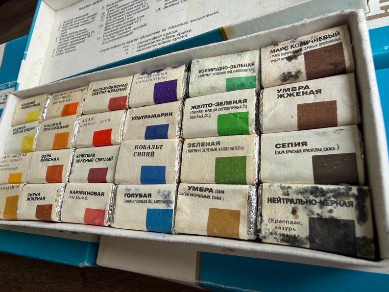 May include: A rectangular white box containing twenty-four individual watercolor paint pans wrapped in paper. Each pan features a color swatch and Russian text labels. The set includes various shades of yellow, orange, red, blue, green, brown, and black.