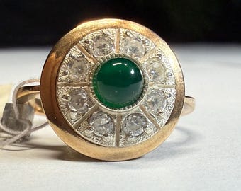 May include: A gold ring featuring a central, round, deep green stone surrounded by a halo of clear, round stones. The ring's band and outer setting are gold-toned, creating a striking contrast with the gemstones.