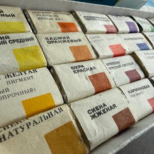 May include: A set of rectangular watercolor paint pans arranged in a cardboard box. Each pan is wrapped in beige paper with black Cyrillic text and a small square color swatch representing the pigment inside, including shades of yellow, orange, red, blue, and brown.
