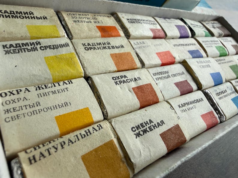 May include: A set of rectangular watercolor paint pans arranged in a cardboard box. Each pan is wrapped in beige paper with black Cyrillic text and a small square color swatch representing the pigment inside, including shades of yellow, orange, red, blue, and brown.