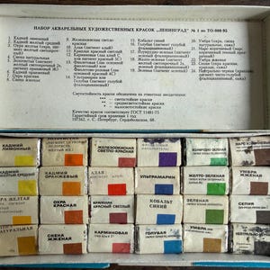 May include: A rectangular white cardboard box containing twenty-four individual watercolor paint pans arranged in four rows of six. Each pan is wrapped in white paper with a color-coded square and Russian text. The lid interior lists the colors.