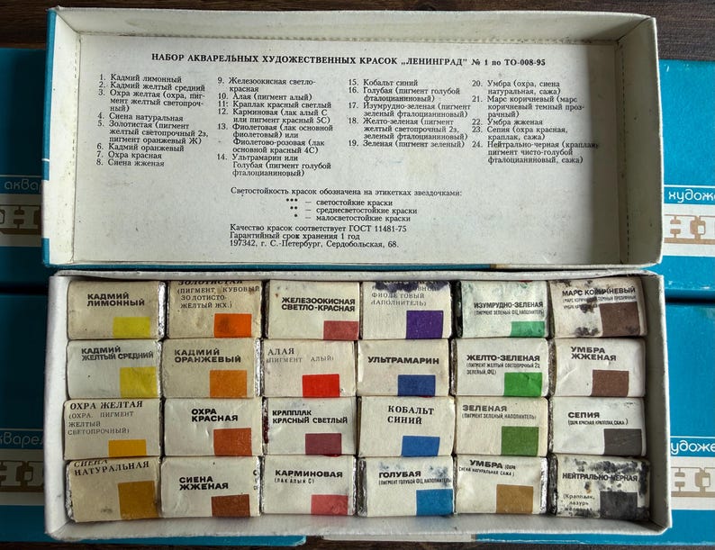 May include: A rectangular white cardboard box containing twenty-four individual watercolor paint pans arranged in four rows of six. Each pan is wrapped in white paper with a color-coded square and Russian text. The lid interior lists the colors.