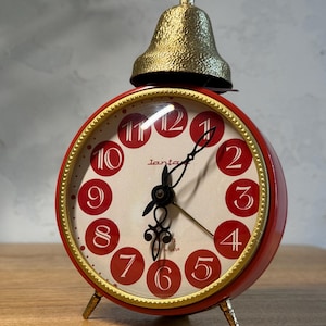 May include: A round red alarm clock with a gold-coloured rim and a textured gold bell on top. The clock face features a cream background with red circles containing white numbers from one to twelve. It has black hands and the text Lanta printed.