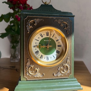 Op de afbeelding: Een rechthoekige groene schoorsteenklok met een donkerbronskleurige wijzerplaat met goudkleurige, sierlijke hoekaccenten. De klok heeft een ronde wijzerplaat met zwarte Romeinse cijfers, een groen midden en gouden wijzers. Bovenop zit een kleine metalen ring.