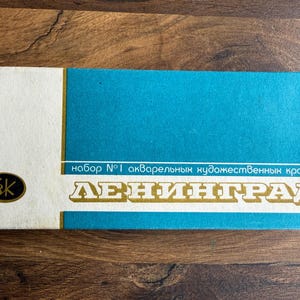 May include: A rectangular watercolor paint set box featuring a white left section with a black logo and a large teal right section. The box displays the text "набор №1 акварельных художественных красок" and the word "ЛЕНИНГРАД" in large white letters.