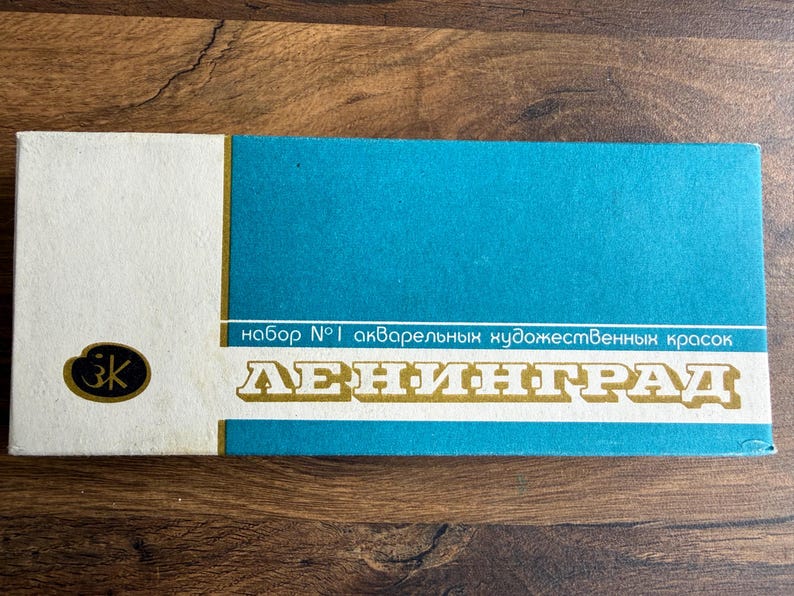 May include: A rectangular watercolor paint set box featuring a white left section with a black logo and a large teal right section. The box displays the text "набор №1 акварельных художественных красок" and the word "ЛЕНИНГРАД" in large white letters.
