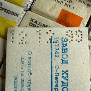 May include: A collection of rectangular paint pans wrapped in off-white paper with Cyrillic text and color swatches. A prominent white paper label features perforated numbers 12 12 99 and blue printed text including ZAVOD KHUDOZHESTVENNYKH KRASOK.