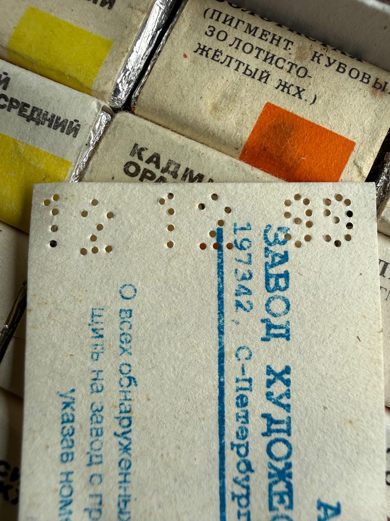 May include: A collection of rectangular paint pans wrapped in off-white paper with Cyrillic text and color swatches. A prominent white paper label features perforated numbers 12 12 99 and blue printed text including ZAVOD KHUDOZHESTVENNYKH KRASOK.