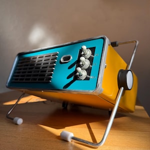 May include: A yellow and teal portable space heater with a rectangular metal body, a front-facing silver grille, and three vertical toggle switches. The device features a silver wire stand with white rubber feet and a side-mounted black control knob.