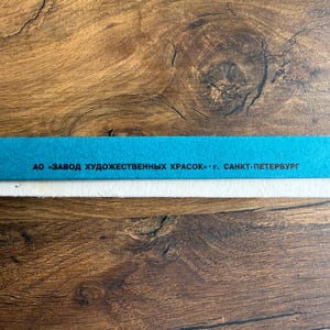 May include: A rectangular box with a bright blue side panel featuring black Cyrillic text that reads AO ZAVOD KHUDOZHESTVENNYKH KRASOK G. SANKT-PETERBURG. The box has a white base and a thin white border along the top edge of the blue section.