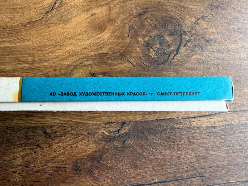 May include: A rectangular box with a bright blue side panel featuring black Cyrillic text that reads AO ZAVOD KHUDOZHESTVENNYKH KRASOK G. SANKT-PETERBURG. The box has a white base and a thin white border along the top edge of the blue section.