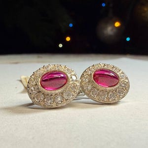 Op de afbeelding: Een paar ovale oorbellen. Elke oorbel heeft een helder roze centrale steen, omringd door een halo van kleinere, heldere edelstenen. De zetting is zilverkleurig, wat zorgt voor een klassieke en elegante uitstraling.