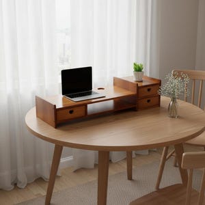 May include: A wooden monitor stand with a laptop, two drawers, and a small shelf. The stand is a warm brown color and sits on a round wooden table. A small plant in a white pot is on the stand.