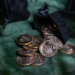 May include: Bronze-coloured coins with embossed designs spilling from a black velvet pouch onto a green surface. The coins feature symbols and patterns, suggesting a fantasy or historical theme.