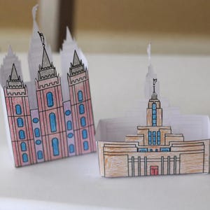 May include: Two papercraft models of religious buildings. One is a pink and blue building with three spires. The other is a tan and blue building with a single spire. Both are hand-drawn and folded.
