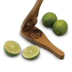 May include: A wooden citrus juicer with a hinged design, featuring a perforated bowl for catching seeds. Two whole green limes and two lime halves are arranged near the juicer, showcasing the product's use. The juicer is made of light brown wood.