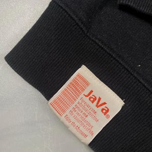 Op de afbeelding: Een zwart kledingstuk van stof met een rechthoekig, gebroken wit label dat aan de zoom is gestikt. Het label toont de rode tekst "JaVa" boven kleinere rode tekst die luidt: "For everyone, who loves pristine nature and the sea and enjoys the adventures of life. Enjoy the moment!"
