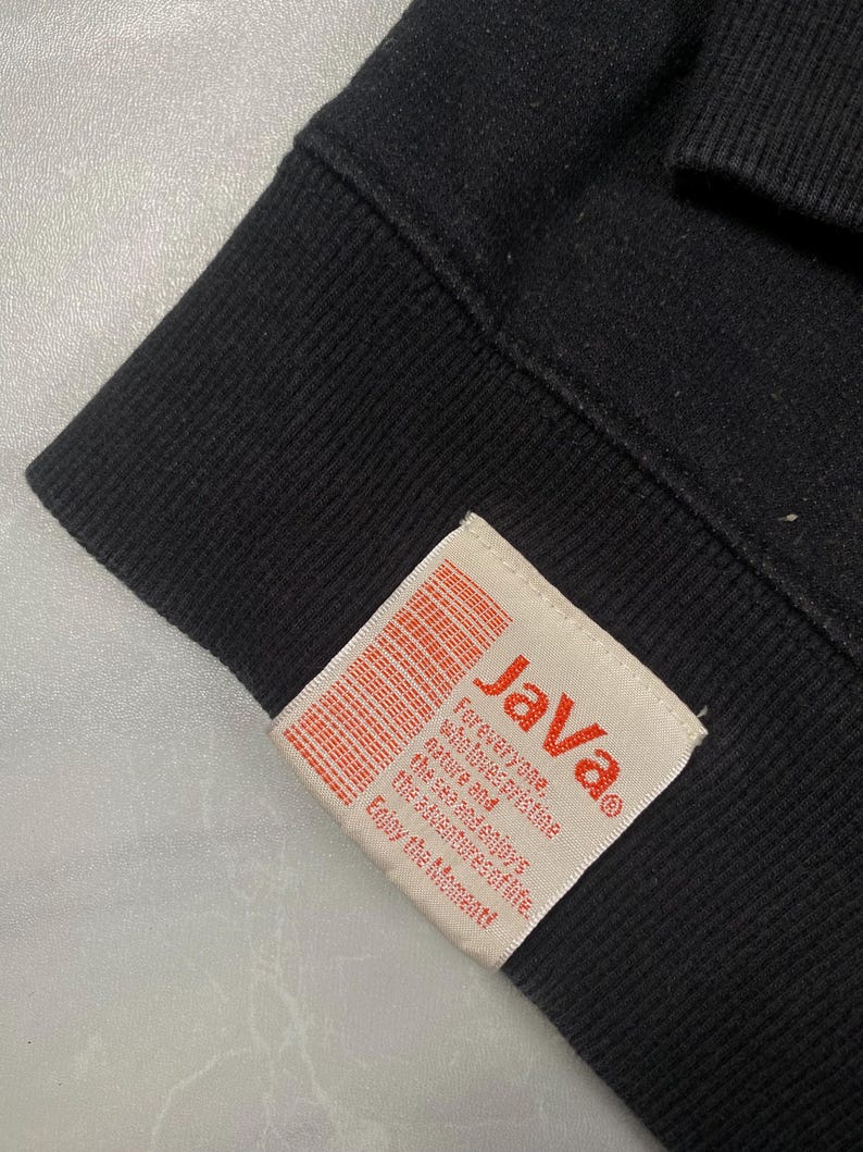 Op de afbeelding: Een zwart kledingstuk van stof met een rechthoekig, gebroken wit label dat aan de zoom is gestikt. Het label toont de rode tekst "JaVa" boven kleinere rode tekst die luidt: "For everyone, who loves pristine nature and the sea and enjoys the adventures of life. Enjoy the moment!"
