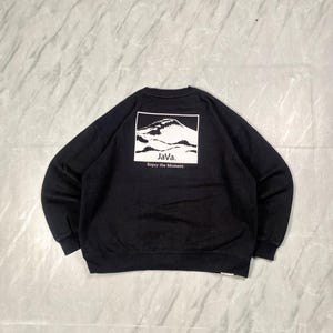 May include: A black long-sleeve sweatshirt featuring a white rectangular graphic on the back. The graphic depicts a mountain range with the text "JaVa." and "Enjoy the Moment." printed in black below the mountain illustration.