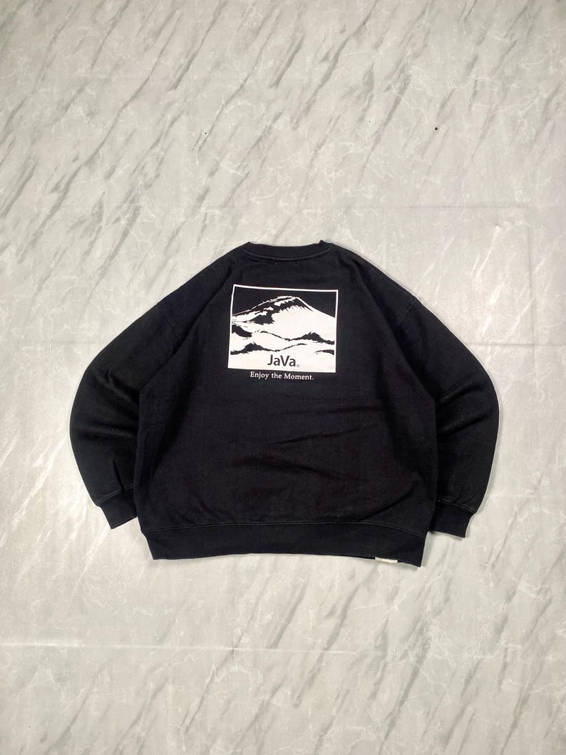 Op de afbeelding: Een zwart sweatshirt met lange mouwen en een witte rechthoekige afbeelding op de achterkant. De afbeelding toont een bergketen met de tekst "JaVa." en "Enjoy the Moment." in het zwart onder de illustratie van de berg.
