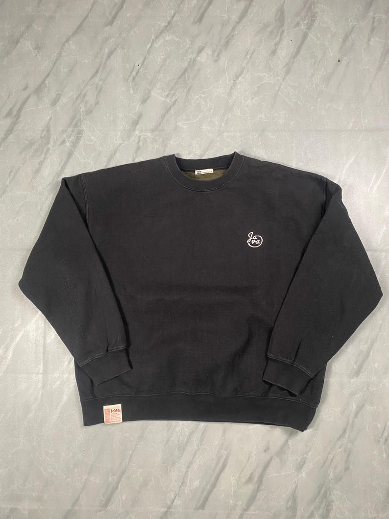 Op de afbeelding: Een zwart sweatshirt met lange mouwen en een ronde hals, voorzien van een klein wit cirkelvormig logo op de linkerborst met de tekst "Java". Een klein rechthoekig wit label met de tekst "Java" is vastgenaaid aan de rechteronderzoom.