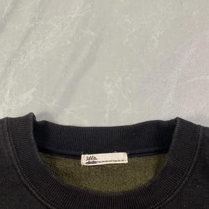 Op de afbeelding: Een zwarte sweater met ronde hals, geribde kraag en een binnenvoering van donker olijfgroen fleece. Een wit rechthoekig stoffen label is in de binnenhals genaaid, met de tekst "Java." en "Enjoy the Moment Enjoy the Life".