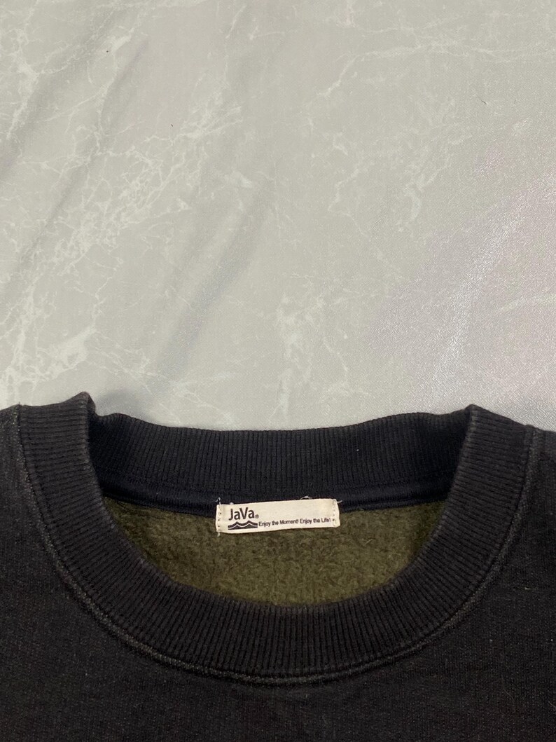 Op de afbeelding: Een zwarte sweater met ronde hals, geribde kraag en een binnenvoering van donker olijfgroen fleece. Een wit rechthoekig stoffen label is in de binnenhals genaaid, met de tekst "Java." en "Enjoy the Moment Enjoy the Life".