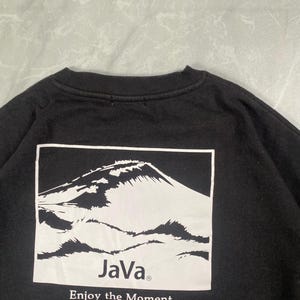 Op de afbeelding: Een zwart sweatshirt met ronde hals en een witte rechthoekige afbeelding op de achterkant. De afbeelding toont een bergtop met de tekst "JaVa" en de zin "Enjoy the Moment." in zwarte letters onder de illustratie van de berg.