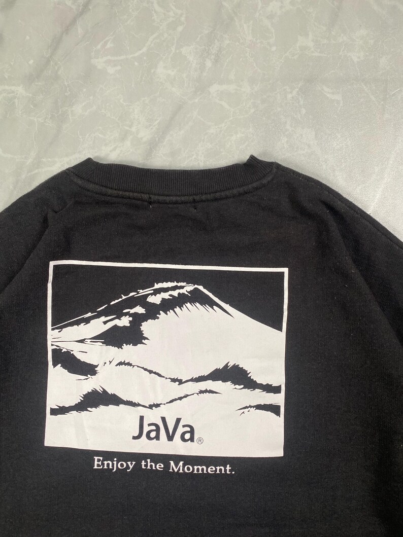 Op de afbeelding: Een zwart sweatshirt met ronde hals en een witte rechthoekige afbeelding op de achterkant. De afbeelding toont een bergtop met de tekst "JaVa" en de zin "Enjoy the Moment." in zwarte letters onder de illustratie van de berg.
