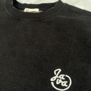 Op de afbeelding: Een zwart sweatshirt met ronde hals, een geribbelde kraag en een klein wit geborduurd logo aan de rechteronderkant. Het logo bestaat uit de cursieve tekst "Java" binnen een cirkelvormige omlijning. De stof heeft een zachte, matte textuur.