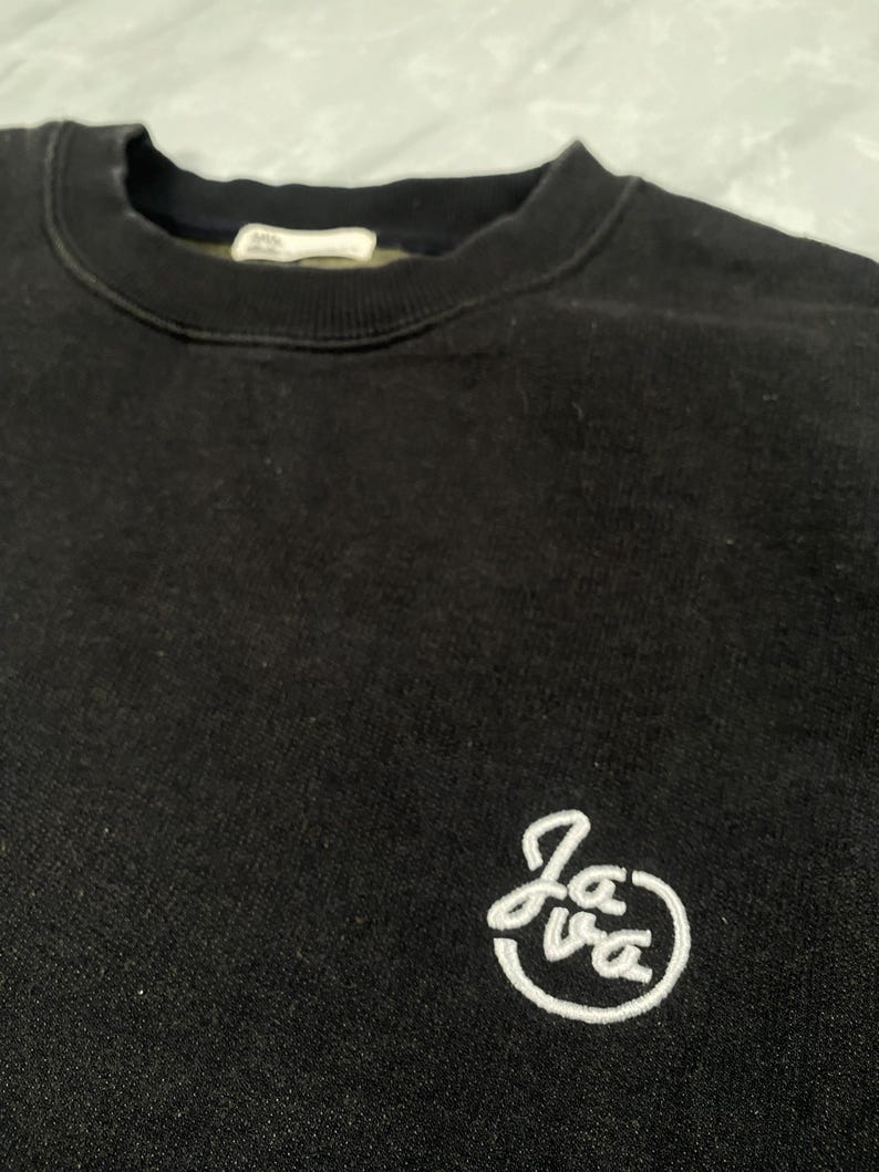 Op de afbeelding: Een zwart sweatshirt met ronde hals, een geribbelde kraag en een klein wit geborduurd logo aan de rechteronderkant. Het logo bestaat uit de cursieve tekst "Java" binnen een cirkelvormige omlijning. De stof heeft een zachte, matte textuur.