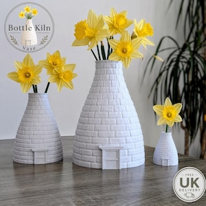 May include: Three grey ceramic kiln-shaped vases, each with a small door, display yellow daffodils. The vases are of different sizes, with the largest in the centre. The vases are on a wooden surface.