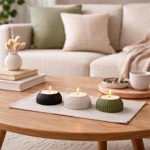 May include: Three lit tealight candles in black, grey, and green holders sit on a beige linen runner on a wooden coffee table. A small vase with dried flowers and books are on the left. A sofa with pillows is in the background.