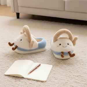 Könnte beinhalten: Ein Paar skurrile, plüschige Hausschuhe in Form von lächelnden Teekannen, mit beigen Griffen und braunen Beinen. Die Hausschuhe sind weiß mit blauen Akzenten. Ein Notizbuch und ein Stift befinden sich im Vordergrund.