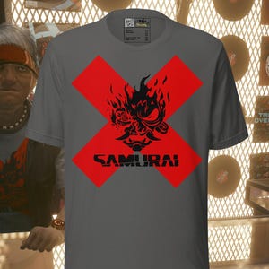 May include: Gray t-shirt featuring a red cross design with a black samurai graphic and the word "SAMURAI" in black. The shirt is displayed in a shop setting with vinyl records and a person in the background.