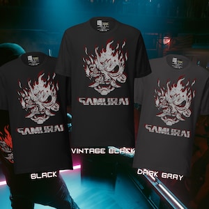 May include: Three short-sleeved t-shirts in black, vintage black, and dark gray. Each shirt features a white and red graphic of a samurai mask with flames and the word "SAMURAI" in white block letters. The shirts are displayed against a blurred background.