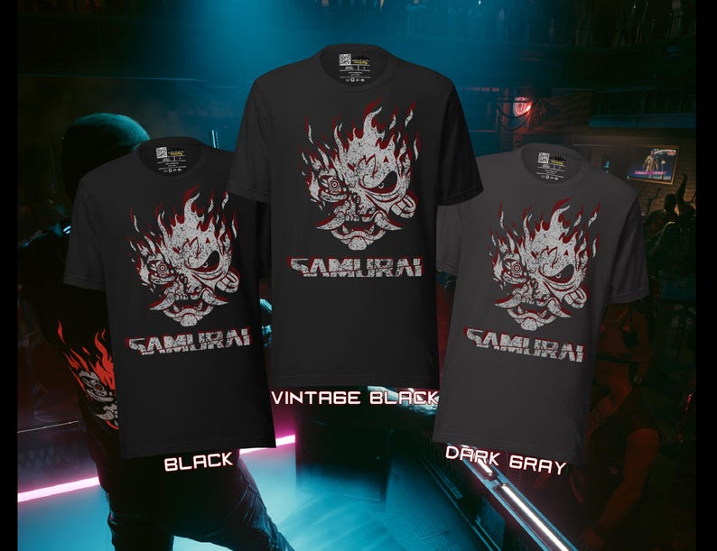 May include: Three short-sleeved t-shirts in black, vintage black, and dark gray. Each shirt features a white and red graphic of a samurai mask with flames and the word "SAMURAI" in white block letters. The shirts are displayed against a blurred background.