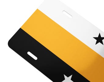 May include: A rectangular luggage tag with a color block design. The tag features a white top section, a gold middle section, and a black bottom section. Two black stars and a white star are visible. The tag has two slots for a strap.