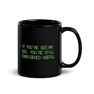 Può includere: Tazza da caffè in ceramica nera con manico curvo. La tazza presenta un testo verde: "IF YOU'RE SEEING ADS, YOU'RE STILL CONSIDERED USEFUL."