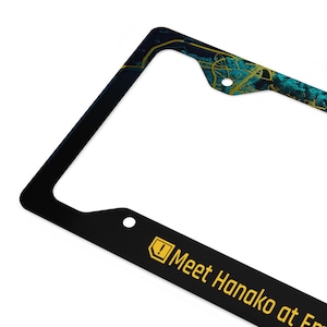May include: Black license plate frame with a teal and gold abstract design in the upper corner. The bottom section has the text "Meet Hanako at Em" in yellow.