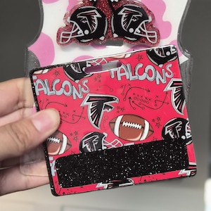 Może przedstawiać: Czerwono-czarny uchwyt na karty w stylu Atlanta Falcons z pasującą kokardą i dwiema przypinkami w kształcie hełmu. Uchwyt na karty ma powtarzający się wzór piłek nożnych, hełmów i logo drużyny. Kokarda ma czarno-biały wzór w szachownicę.