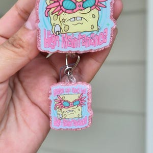 May include: Two acrylic badge reels featuring a cartoon Spongebob Squarepants wearing pink glasses. The text on the badges reads "Wake up and be High Maintenance" in pink lettering. The badges are attached to a clear reel.
