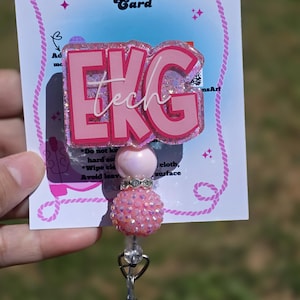 Può includere: Un portabadge EKG tech rosa e glitterato. Il portabadge presenta il testo "EKG tech" in lettere rosa. Il portabadge è attaccato a una carta bianca con il testo "Badge Reel Care Card".