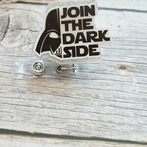Könnte beinhalten: Ein silberner Glitzer-Badge-Roller mit Darth Vaders Helm und dem Text "JOIN THE DARK SIDE" in Schwarz und Weiß. Der Badge-Roller hat einen transparenten Kunststoffriemen und einen Metallclip.