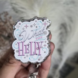 May include: A white badge reel with the text "What the HELLY" in pink and black script. The badge has a glittery silver border and is decorated with stars and swirls. A clear plastic strap and metal clip are attached.